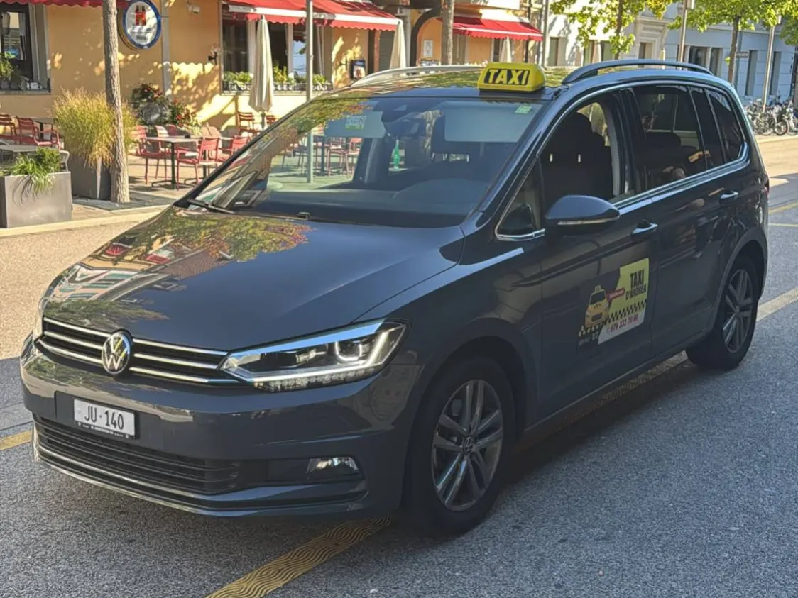 Taxi d'Andrea VW Touran gris JU 140 devant une terrasse du centre de Delémont, service taxi 24h/24 dans le Jura